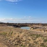 A dirt path runs through dry, grassy land along Jones Rd toward a small, calm river under a mostly clear blue sky with a few scattered clouds in Woodson, TX 76491.