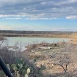 A desert landscape near Woodson TX with prickly pear cacti, dry brush, and a small body of water under a partly cloudy sky. The edge of a green vehicle is visible in the foreground, hinting at travel along 1502 Jones Rd.