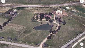 Aerial view of a rural Aledo, TX property at 2301 FM 1187 with a large house, several smaller buildings, a pond surrounded by trees, fenced areas, and adjacent roads and fields.