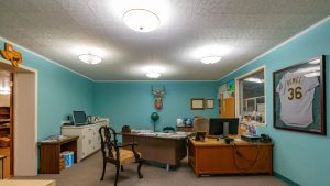 A teal-walled office with two desks, computers, a vintage chair, wall-mounted deer head, and a framed sports jersey. The room is well-lit with ceiling lights and decorated with photos and certificates.