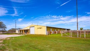 A large yellow barn-like building with a white garage door sits on a grassy field, surrounded by wire fencing under a bright blue sky with scattered clouds and contrails.