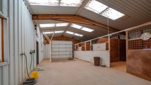 A clean, spacious horse stable with metal and wood stalls, concrete floor, a hose, bucket, trash can, and overhead skylights letting in natural light. A large door is at the end of the corridor.