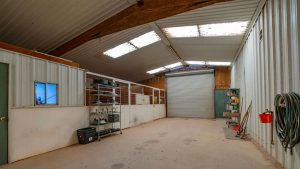 A spacious, well-lit barn or workshop with a high, sloped metal roof, white walls, shelves with tools and supplies, a large rolling door, and a coiled hose hanging on the wall.