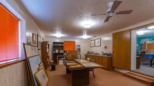 An office room with wood panel walls, framed maps leaning against a table, cardboard boxes, bookshelves, cabinets, and orange blinds covering two windows. An open door leads to another room with a bulletin board.