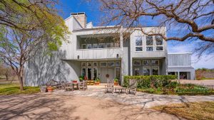Modern two-story house with large windows and a gray exterior, surrounded by leafless trees. A stone patio in front features wooden chairs, tables, and potted plants under a clear blue sky.