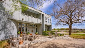 Modern two-story house with gray siding, large windows, and a spacious stone patio featuring outdoor wooden furniture and potted plants. Leafy trees surround the yard, and the sky is mostly clear.