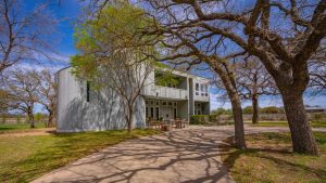 A modern, two-story white house with large windows and a curved facade is surrounded by trees casting shadows on the driveway and lawn under a bright blue sky. Patio furniture is set near the entrance.