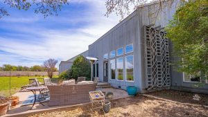 Modern gray house with large windows and a spacious patio featuring outdoor seating, a dining table, and potted plants, surrounded by grass, trees, and open sky on a sunny day.