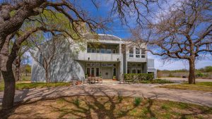 A modern two-story gray house with large windows and a balcony, surrounded by leafless trees. Outdoor furniture sits on a patio in front, and shadows of branches stretch across the yard and walkway.
