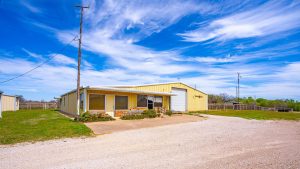 A yellow metal building with a covered porch sits on a gravel driveway under a bright blue sky with wispy clouds, surrounded by grass and open land. A utility pole stands nearby.