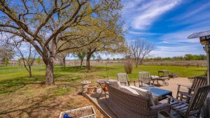 A backyard patio with outdoor seating, including chairs and a sofa, overlooks a spacious grassy yard with large trees and a fence in the distance under a bright, blue sky with scattered clouds.