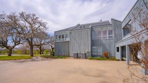 A modern two-story gray house with large windows, surrounded by leafless trees and greenery, sits next to a spacious concrete driveway under a partly cloudy sky.