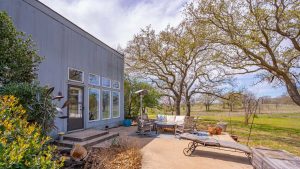 A modern gray house with large windows opens to a patio with outdoor seating, a lounge chair, potted plants, and a view of grassy fields and leafless trees under a partly cloudy sky.