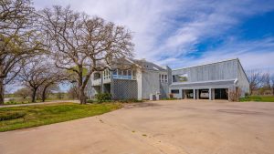 A modern, gray, two-story house with large windows and geometric design sits next to leafless trees, with a spacious concrete driveway and a bright blue sky with wispy clouds above.