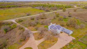 Aerial view of a large house with a gray roof, surrounded by trees and open fields. Curved driveway and road nearby. Rural landscape with green grass and scattered buildings in the distance.