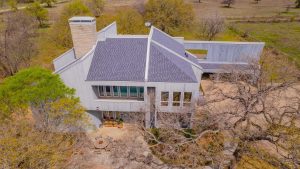 Aerial view of a modern two-story house with gray siding, large windows, and an angled roof, surrounded by trees and a stone patio in a rural landscape.