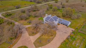 Aerial view of a large house with a gray roof, circular driveway, three-car garage, and surrounding trees on a spacious, partially wooded property near a rural road.