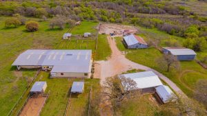 Aerial view of a rural property with several barns and sheds, fenced horse paddocks, dirt roads, and green fields surrounded by trees and vegetation.
