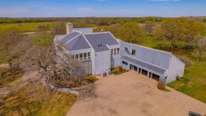 Aerial view of a modern, light gray house with geometric architecture, large windows, and a spacious driveway, surrounded by trees and open grassy fields under a blue sky.
