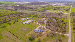 Aerial view of a rural property with several buildings, open fields, trees, and winding dirt roads, surrounded by greenery and adjacent to a long, straight road; distant hills and a town visible in the background.