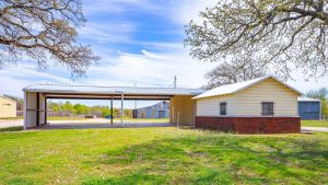 A small cream-colored building with a metal roof and red brick base sits beside a covered carport on a grassy lawn, with trees and additional outbuildings visible in the background under a blue sky.