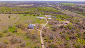Aerial view of a rural property with scattered buildings, winding roads, patches of trees, and open grassy fields, surrounded by wooded areas and rolling hills under a partly cloudy sky.