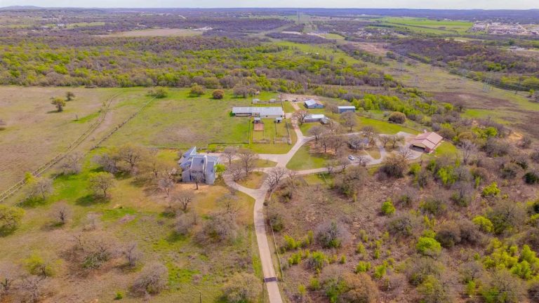 Aerial view of a rural property with scattered buildings, winding roads, patches of trees, and open grassy fields, surrounded by wooded areas and rolling hills under a partly cloudy sky.