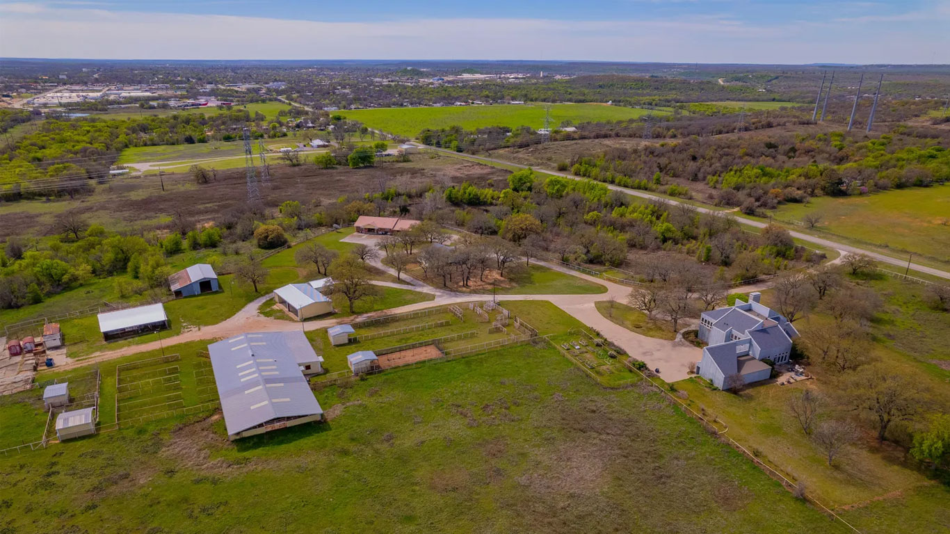 Aerial view of a rural property with several farm buildings, fenced areas, pastures, and trees, surrounded by green fields and distant cityscape under a partly cloudy sky.
