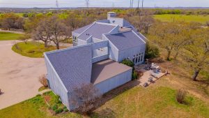 A modern, angular house with gray siding and multiple sloped roofs sits among trees and grassy fields, featuring an outdoor patio area with seating and nearby paved driveway.