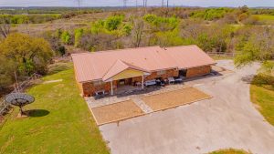 Aerial view of a single-story brick house with a metal roof, surrounded by trees and grass, a large paved driveway, and a satellite dish in the yard. Power lines and hills are visible in the background.