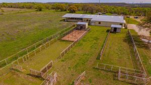 Aerial view of a rural property with fenced animal pens, grassy areas, and a large metal barn surrounded by open fields and scattered trees under a clear sky.