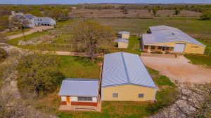 Aerial view of a rural property with multiple buildings, including a small white house and two large yellow metal barns, surrounded by fenced grassy areas, trees, and open fields under a blue sky.
