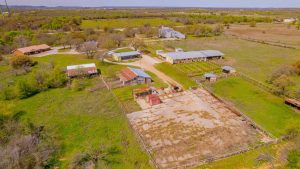 Aerial view of a rural ranch with multiple barns, fenced areas, animal pens, trees, and open grassy fields under a clear sky. Several buildings and a main house are visible among the landscape.