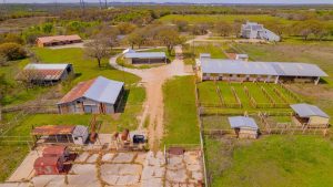 Aerial view of a ranch with fenced horse pens, several barns and sheds, a dirt road, grassy fields, and scattered trees. Farm buildings and a large white house are visible in the background.