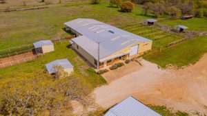 Aerial view of a large metal barn with a gray roof, surrounded by fenced grassy areas, smaller sheds, and a dirt driveway in a rural setting. Trees and open fields are visible in the background.