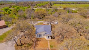 Aerial view of a small light-colored house with a gray roof surrounded by leafless trees and brown grass, situated near a road with green fields and hills in the background.