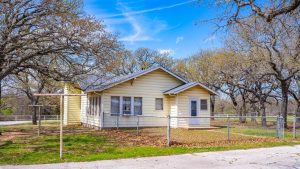 A small yellow house with a gray roof is surrounded by a chain-link fence and bare trees. Dry leaves cover the ground, and the sky is bright blue with wispy clouds.