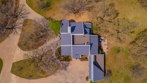 Aerial view of a house with a gray roof surrounded by leafless trees, winding driveways, and patches of grass in a spacious yard.