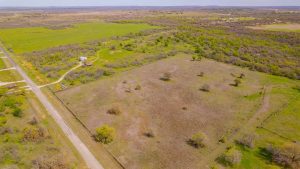 Aerial view of a rural landscape featuring a large, fenced grassy field with scattered trees, a dirt road running along one edge, and several houses and outbuildings surrounded by green fields and wooded areas.