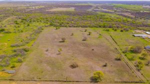 Aerial view of a large, fenced grassy field with scattered trees, surrounded by more green fields, trees, and distant buildings under a clear sky.