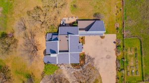 Aerial view of a house with a gray roof, surrounded by dry grass, trees, a large driveway, and a garden with rectangular plots on the right side.