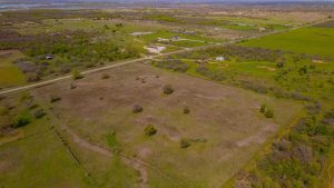 Aerial view of a rural landscape with green fields, scattered trees, fenced areas, dirt roads, and a few houses, with distant hills and a river visible in the background.