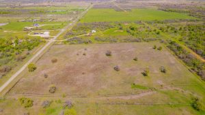 Aerial view of a rural landscape with a large fenced field, scattered trees, green farmland, houses, and long roads stretching through the countryside under a clear sky.