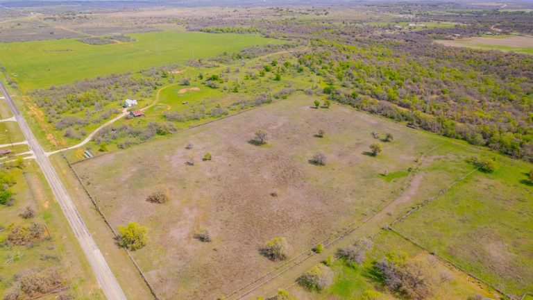 Aerial view of a large, rural, grassy property divided by fences, with scattered trees, a nearby house, a road along the edge, and surrounding fields and woodlands in the background.