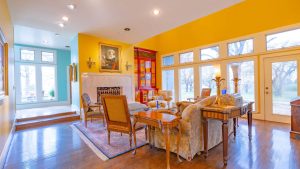 A bright, elegant living room with yellow walls, large windows, classic furniture, a fireplace with a portrait above it, a red display cabinet, and hardwood floors with an area rug. Natural light fills the space.