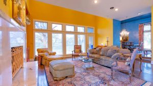 Bright living room with yellow walls, large windows, ornate furniture, a patterned rug, and a marble fireplace. A dining area with a chandelier is visible in the background.