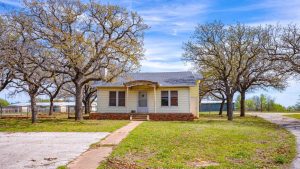 A small, single-story yellow house with a dark roof sits between large leafless trees. A concrete path leads to the front door, surrounded by a grassy yard under a blue sky with light clouds.