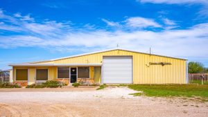 A yellow metal building with a large white garage door, several windows, and a sign reading "STARS & STRIPES BARN" under a clear blue sky, with gravel and grass in the foreground.