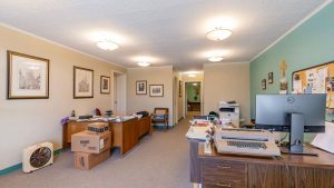 A spacious office with two desks, scattered papers, computer monitors, cardboard boxes, framed art on beige and green walls, bulletin boards, and overhead lighting. The room is tidy but busy, with some office equipment and supplies visible.
