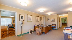 A well-lit office with beige walls, desks with stacks of papers and boxes, framed portraits on the wall, and a hallway visible in the background. A hand sanitizer dispenser stands near the entrance.
